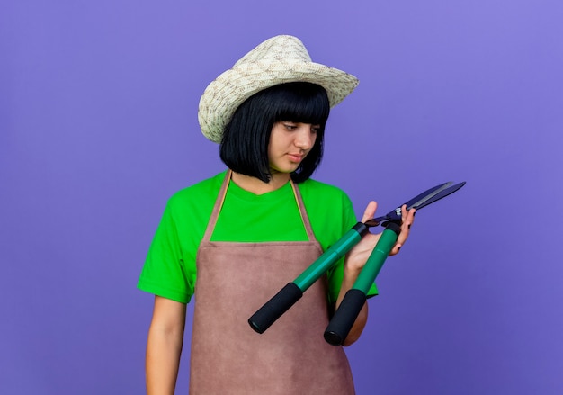 Selbstbewusste junge Gärtnerin in Uniform mit Gartenhut hält Schermaschinen