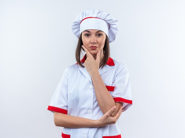Selbstbewusste junge Köchin in Kochuniform, die Hand auf das Kinn legt, isoliert auf weißer Wand