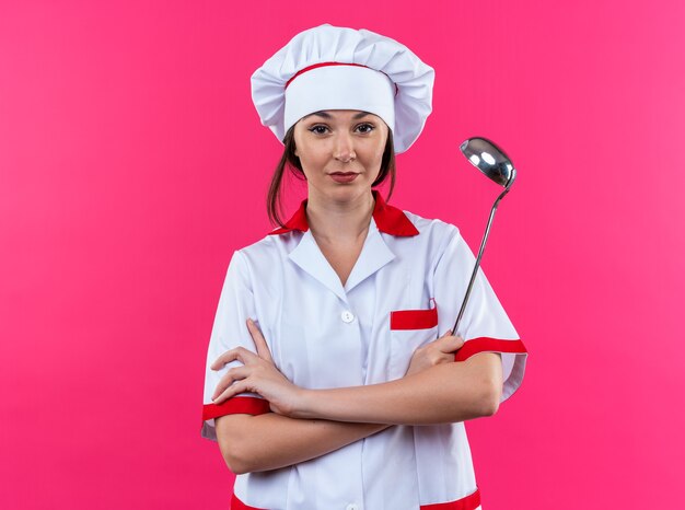 selbstbewusste junge Köchin mit Kochuniform, die die Hände überquert, isoliert auf rosa Wand