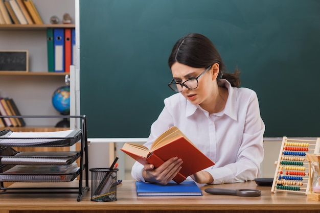 Selbstbewusste junge Lehrerin, die ein Buch hält und liest, das am Schreibtisch mit Schulwerkzeugen im Klassenzimmer sitzt