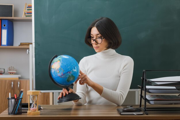 Selbstbewusste junge Lehrerin, die eine Brille trägt und auf die Weltkugel zeigt, die am Schreibtisch mit Schulwerkzeugen im Klassenzimmer sitzt