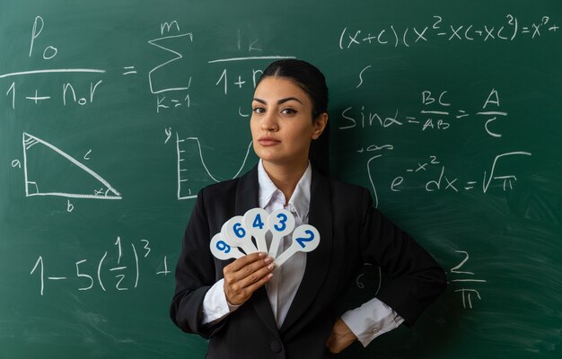 selbstbewusste junge Lehrerin, die vor der Tafel steht und Zahlenfans hält, die im Klassenzimmer die Hand an Bord legen