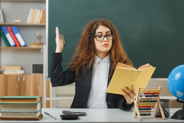 Selbstbewusste junge Lehrerin mit Brille, die ein Buch hält und liest, das am Schreibtisch mit Schulwerkzeugen im Klassenzimmer sitzt