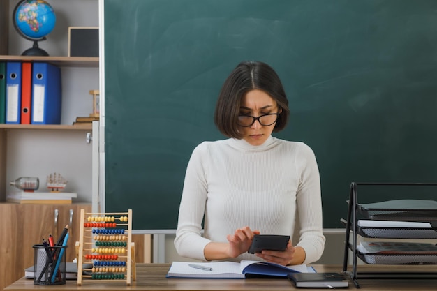Selbstbewusste junge Lehrerin mit Brille, die einen Taschenrechner hält und betrachtet, der am Schreibtisch mit Schulwerkzeugen im Klassenzimmer sitzt