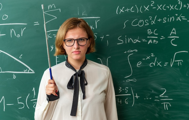 Selbstbewusste junge Lehrerin mit Brille, die vor der Tafel steht und einen Zeigerstock im Klassenzimmer hält holding