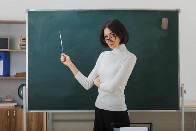 Selbstbewusste junge Lehrerin mit Brille, die vor der Tafel steht und mit einem Zeiger im Klassenzimmer auf die Tafel zeigt