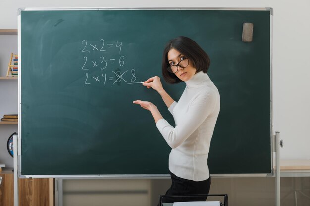 Selbstbewusste junge Lehrerin mit Brille steht vor der Tafel und schreibt im Klassenzimmer