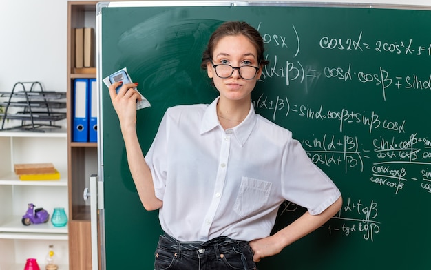 Kostenloses Foto selbstbewusste junge mathematiklehrerin mit brille, die vor der tafel steht und die hand auf der taille hält, die tafelradierer hält und nach vorne im klassenzimmer schaut