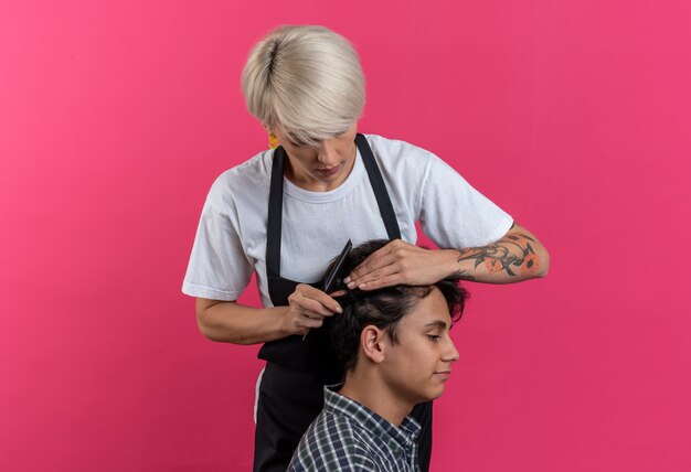 Selbstbewusste junge schöne Friseurin in Uniform, die Haarschnitt für Jungen macht, isoliert auf rosa Wand