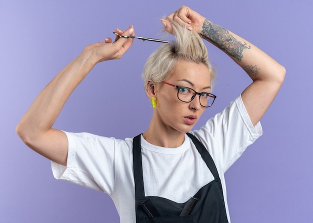 Selbstbewusste junge schöne Friseurin in Uniform mit Brille, die Haare mit einer Schere schneidet, isoliert auf blauer Wand