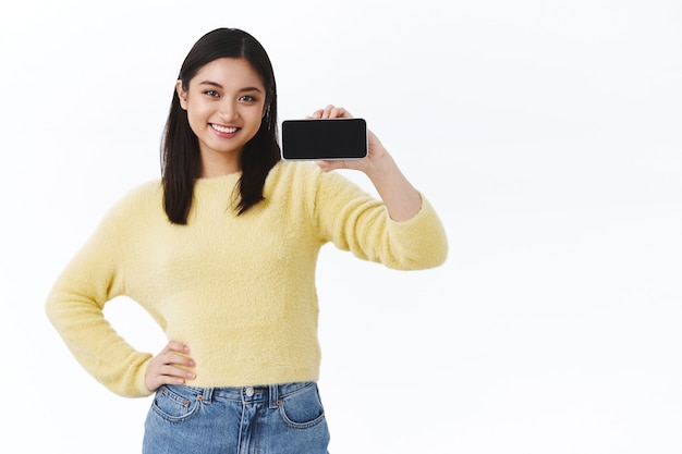 Selbstbewusste junge, süße asiatische Programmiererin, die stolz ihre neue Anwendung zeigt, Smartphone horizontal hält, App oder Spiel auf dem mobilen Bildschirm fördert und erfreut über weiße Wand lächelt