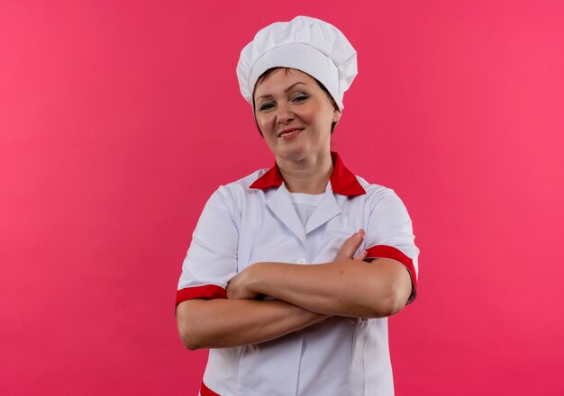 Selbstbewusste Köchin mittleren Alters in Kochuniform, die Hände auf isolierter rosa Wand mit Kopienraum kreuzt