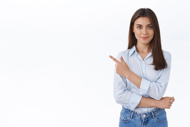 Selbstbewusste, professionelle junge Frau in blauer Bluse, die mit dem Finger in die obere linke Ecke zeigt und in die Kamera schaut, überzeugt den Kunden, die richtige Wahl zu treffen und einen Vertrag mit ihrem weißen Firmenhintergrund zu unterzeichnen