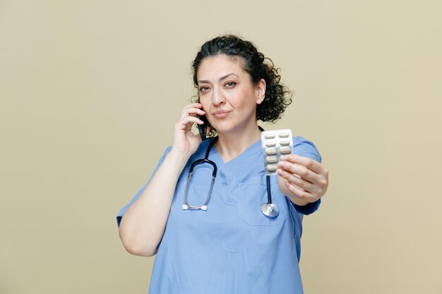 Selbstbewusste Ärztin mittleren Alters, die Uniform und Stethoskop um den Hals trägt und in die Kamera blickt, die eine Packung Pillen in Richtung Kamera streckt, während sie am Telefon isoliert auf olivfarbenem Hintergrund spricht