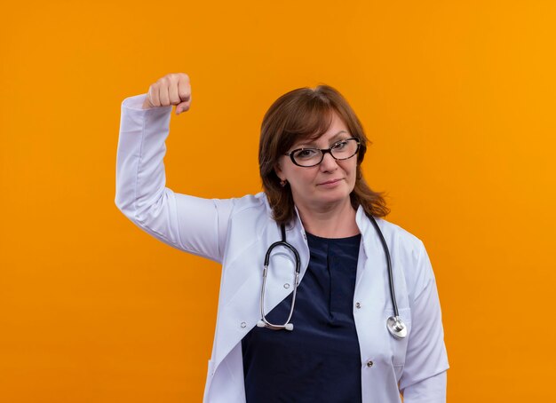 Selbstbewusste Ärztin mittleren Alters mit Brille, medizinischem Gewand und Stethoskop, die die Faust an der isolierten orangefarbenen Wand hebt