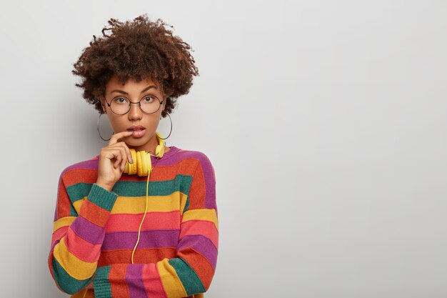 selbstbewusste schöne Frau mit natürlichem lockigem Haar, hält Finger in der Nähe der Lippen, trägt runde Ohrringe, Brille und gestreiften Pullover, Modelle gegen weiße Wand