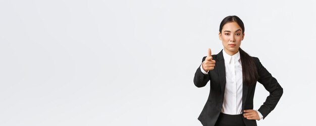 Selbstbewusste, strenge asiatische Unternehmerin, die mit dem Finger auf die Kamera zeigt und ernst aussieht, schlägt ein Stellenangebot vor, um einen Mitarbeiter für die Arbeit mit weißem Hintergrund auszuwählen