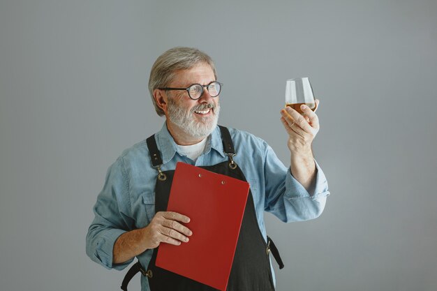 Selbstbewusster älterer Mannbrauer mit selbst hergestelltem Bier im Glas auf Holzfass auf Grau