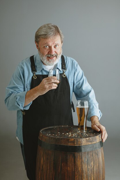 Selbstbewusster älterer Mannbrauer mit selbst hergestelltem Bier im Glas auf Holzfass auf grauer Wand