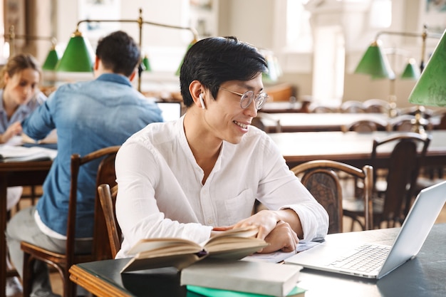 Selbstbewusster asiatischer student mit kopfhörern, der in der bibliothek mit laptop und büchern studiert?