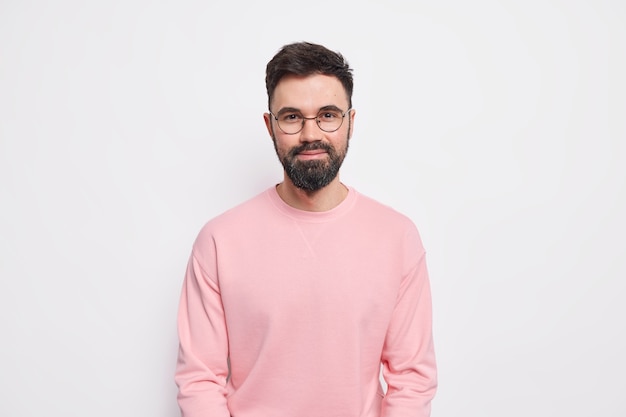 Selbstbewusster bärtiger Macho-Mann sieht zufrieden aus, hat ein freundliches, freundliches Grinsen im Gesicht, trägt eine runde Brille, einen rosa Pullover jump