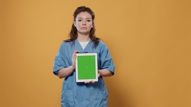 Selbstbewusster Caucasion-Arzt posiert mit digitalem Tablet mit Green-Screen-Mockup-Design in Krankenhausuniform im Studio. Ärztin mit Touchscreen-Gerät zur Werbung für medizinische Konzepte.