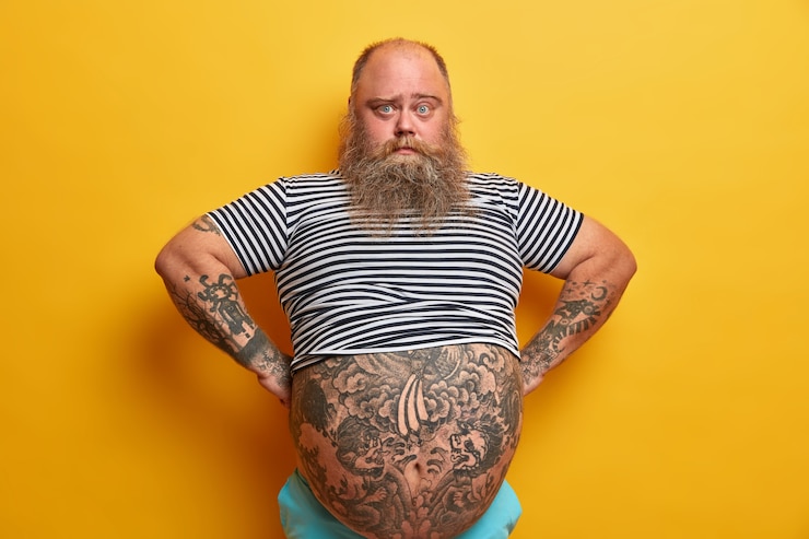Kostenlose Foto selbstbewusster ernsthafter blauäugiger mann mit bart, hat großen bauch, führt ungesunden lebensstil, gekleidet in gestreiftem, untergroßem matrosen-t-shirt, posiert über gelber wand. praller typ steht selbstbewusst drinnen