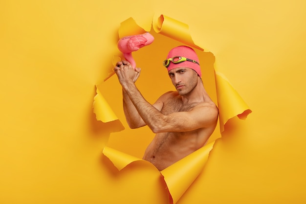 Selbstbewusster ernsthafter Kerl mit Borsten hält rosa aufgeblasenen Flamingo, trägt Badekappe mit Schutzbrille auf der Stirn