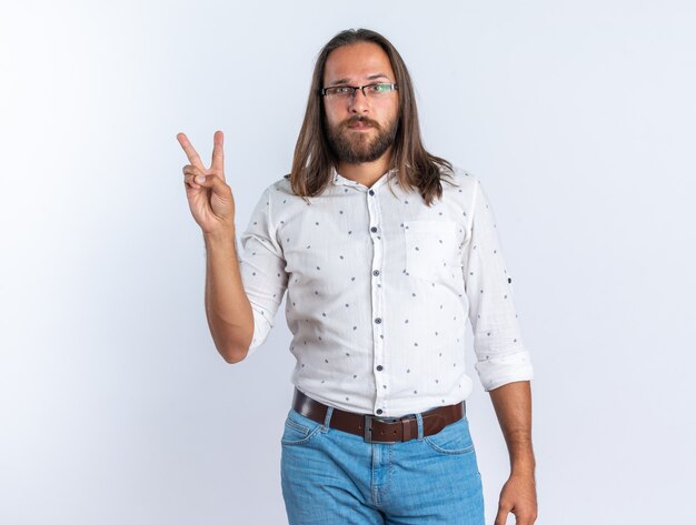 Selbstbewusster erwachsener gutaussehender Mann mit Brille, der die Kamera anschaut und das Friedenszeichen isoliert auf weißer Wand tut