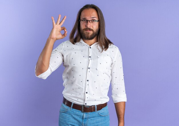 Selbstbewusster erwachsener, gutaussehender Mann mit Brille, der die Kamera anschaut und das OK-Zeichen isoliert auf lila Wand mit Kopienraum tut