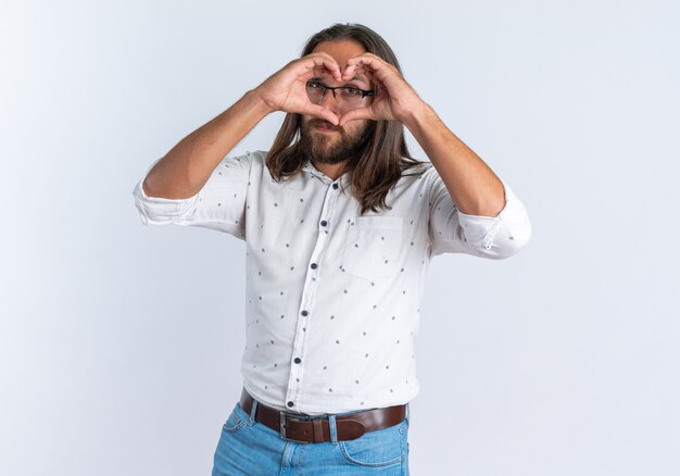 Selbstbewusster erwachsener gutaussehender Mann mit Brille, der die Kamera anschaut und Liebeszeichen vor den Augen isoliert auf weißer Wand macht