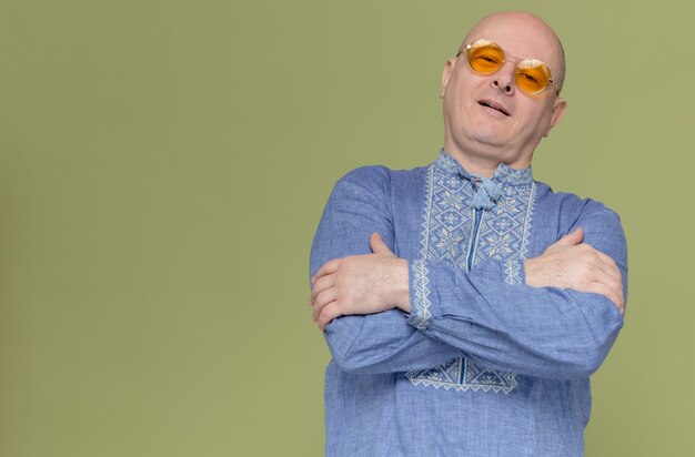 Selbstbewusster erwachsener slawischer Mann im blauen Hemd mit Sonnenbrille, der mit verschränkten Armen steht