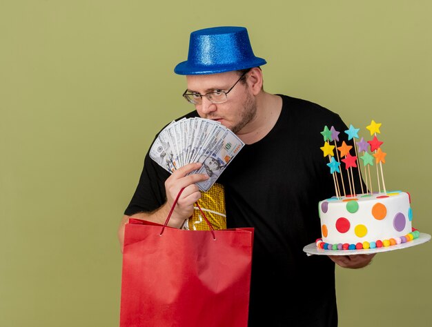 Selbstbewusster erwachsener slawischer Mann in optischer Brille mit blauem Partyhut hält Geldgeschenkbox-Papiereinkaufstasche und Geburtstagstorte