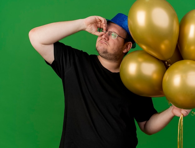 Selbstbewusster erwachsener slawischer Mann in optischer Brille mit blauem Partyhut hält Heliumballons und sieht die Seite durch die Finger an