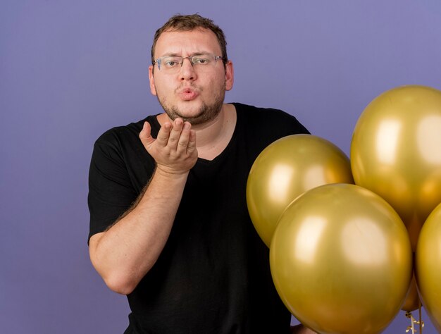 Selbstbewusster erwachsener slawischer Mann in optischer Brille steht mit Heliumballons und sendet einen Kuss mit der Hand