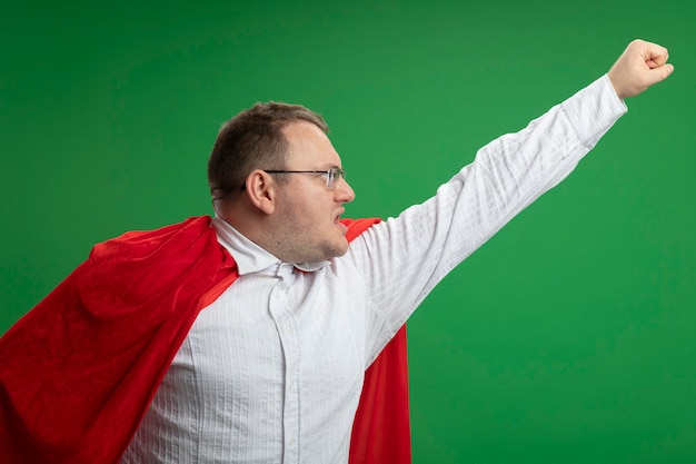 Selbstbewusster erwachsener slawischer Superheldenmann im roten Umhang, der die Brille trägt, die Seite betrachtet, die Faust oben auf grüner Wand anhebt