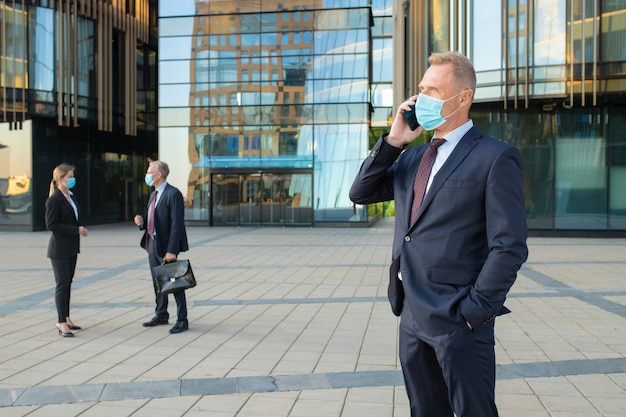 Selbstbewusster Geschäftsmann, der Maske und Büroanzug trägt, die auf Handy draußen sprechen. Geschäftsleute und Stadtbauglasfassade im Hintergrund. Speicherplatz kopieren. Geschäfts- und Epidemiekonzept