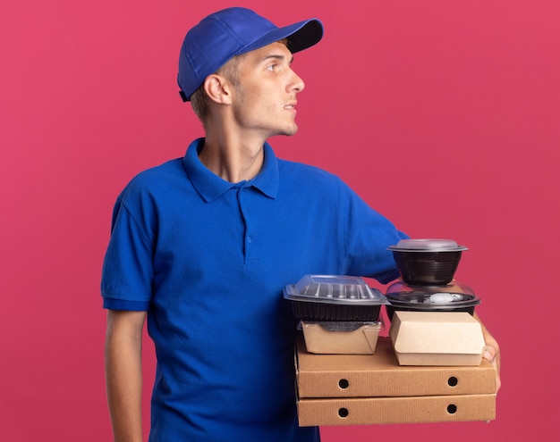 Selbstbewusster junger blonder Lieferjunge hält Lebensmittelbehälter und Pakete auf Pizzakartons mit Blick auf die Seite