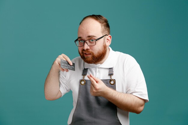 Selbstbewusster junger Friseur in Uniform und Brille mit Kreditkarte, der in die Kamera blickt und Geldgeste isoliert auf blauem Hintergrund zeigt