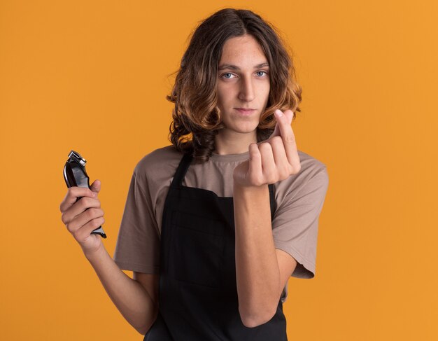 Selbstbewusster junger gutaussehender Friseur in Uniform, der Haarschneidemaschinen hält, die Geldgeste machen