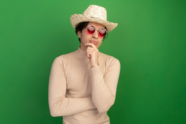 Selbstbewusster junger gutaussehender Kerl mit Hut mit Brille packte das Kinn isoliert auf grüner Wand mit Kopierraum