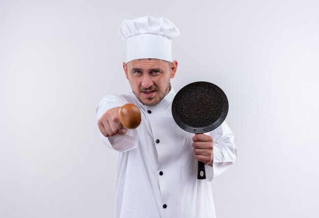 Selbstbewusster junger gutaussehender Koch in Kochuniform, der Bratpfanne hält und mit Löffel auf isolierte weiße Wand zeigt