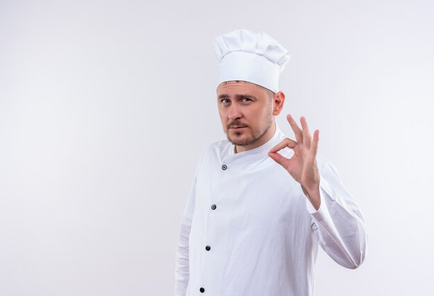 Selbstbewusster junger, gutaussehender Koch in Kochuniform, der das OK-Zeichen isoliert auf weißer Wand mit Kopierraum tut