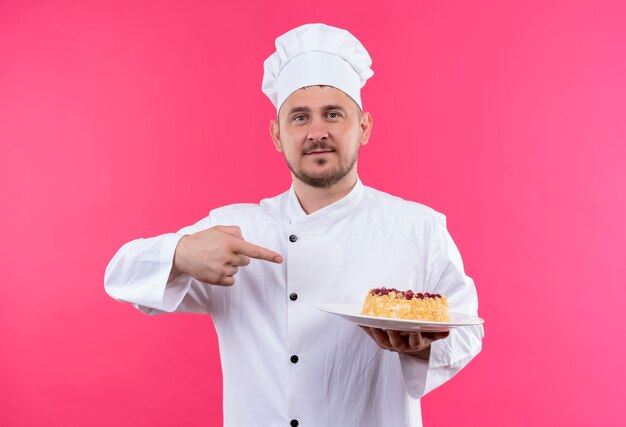 Selbstbewusster junger gutaussehender Koch in Kochuniform, der einen Teller mit Kuchen hält, der darauf isoliert auf rosa Wand zeigt?