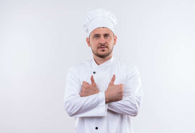 Selbstbewusster junger gutaussehender Koch in Kochuniform, der mit geschlossener Haltung steht und Daumen nach oben zeigt, isoliert auf weißer Wand