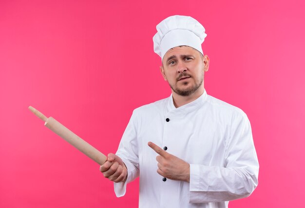 Selbstbewusster junger gutaussehender Koch in Kochuniform, der Nudelholz hält, das auf die Seite zeigt, isoliert auf rosa Wand