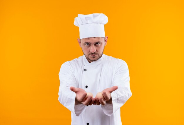 Selbstbewusster junger gutaussehender Koch in Kochuniform, der sich ausstreckt und leere Hände isoliert auf oranger Wand zeigt