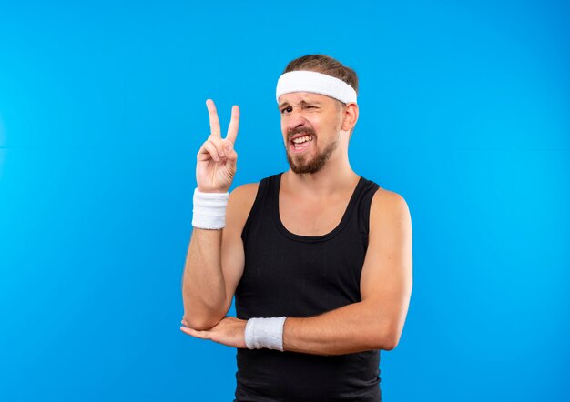 Selbstbewusster junger, gutaussehender, sportlicher Mann mit Stirnband und Armbändern, der ein Friedenszeichen macht und die Hand unter den Ellbogen zwinkert, isoliert auf blauer Wand mit Kopierraum
