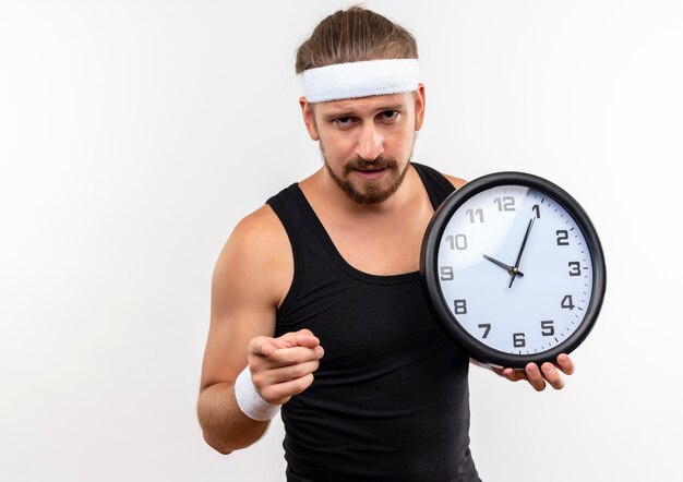 Selbstbewusster junger, gutaussehender, sportlicher Mann mit Stirnband und Armbändern, der eine Uhr hält und isoliert auf eine weiße Wand mit Kopienraum zeigt