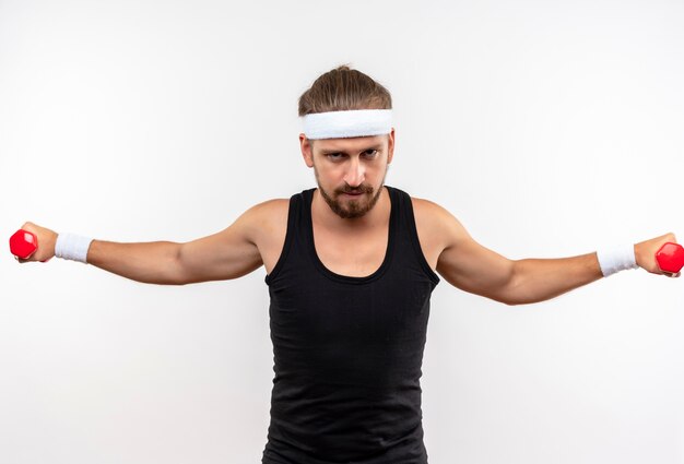 Selbstbewusster junger, gutaussehender, sportlicher Mann mit Stirnband und Armbändern, der Hanteln mit offenen Armen isoliert auf weißer Wand hält und ausstreckt